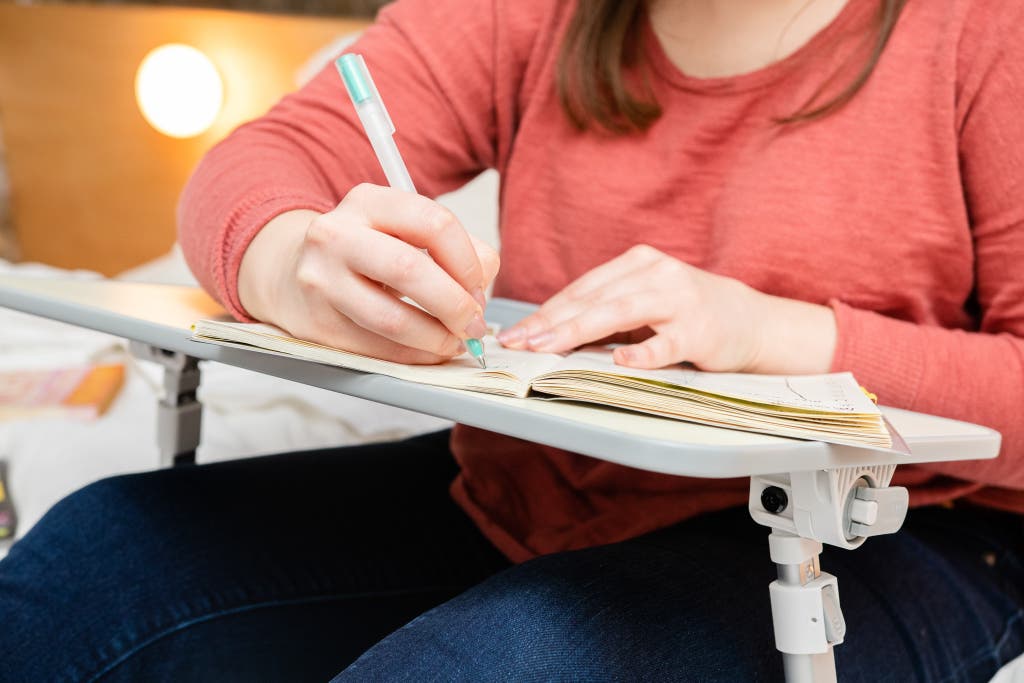 A person seated comfortably, writing on a notepad resting on a white lap desk with legs.